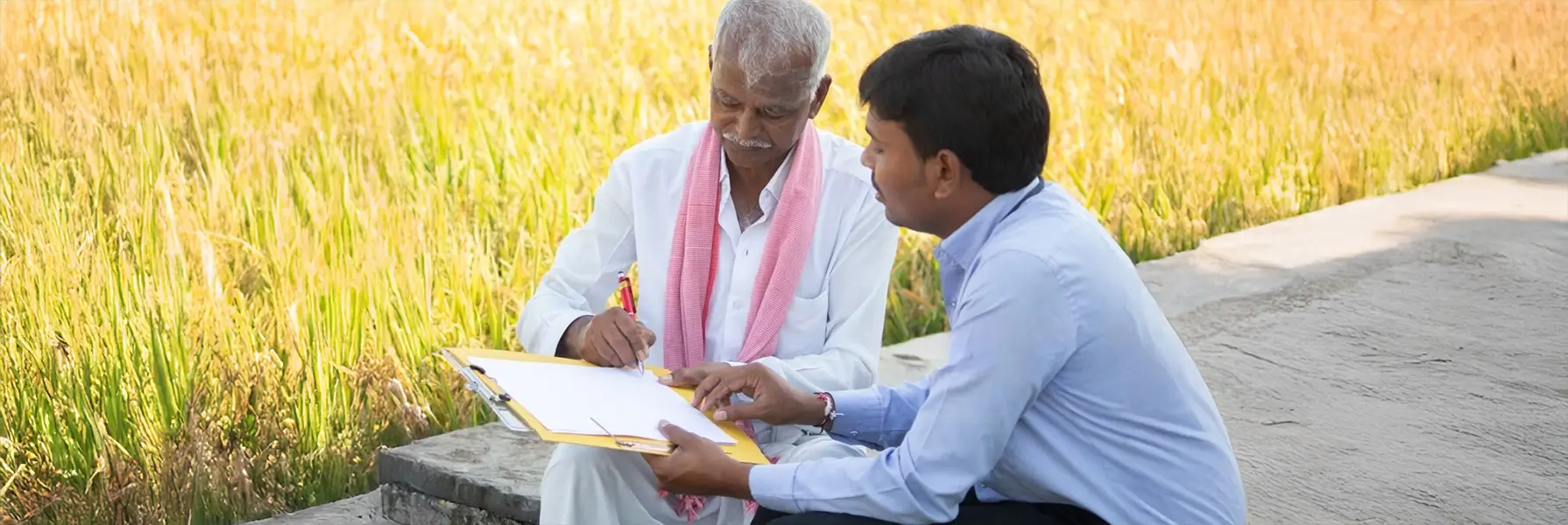 A young man helps a farmer fill out paperwork on a clipboard, sitting near a golden field.